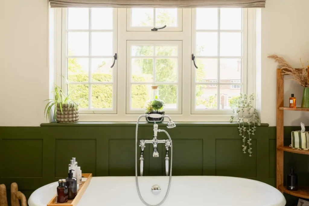 Freestanding bath beneath a window with dark green panelled wall