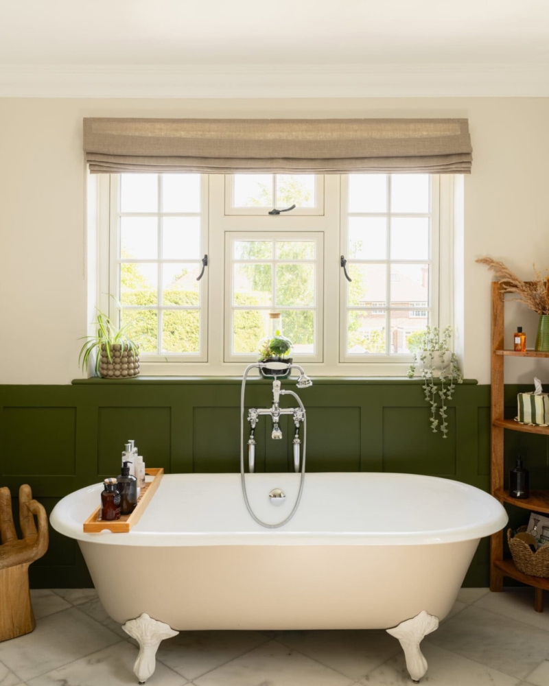 Freestanding bath beneath a window with dark green panelled wall