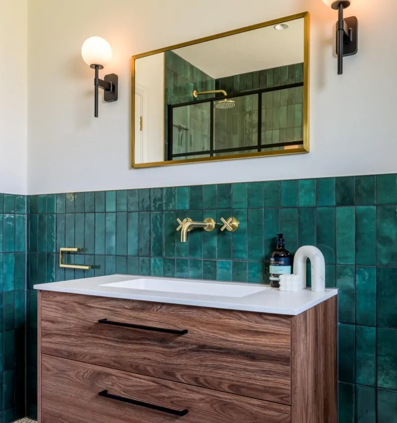 Bathroom vanity with deep green tiles, brass taps and black framed mirror
