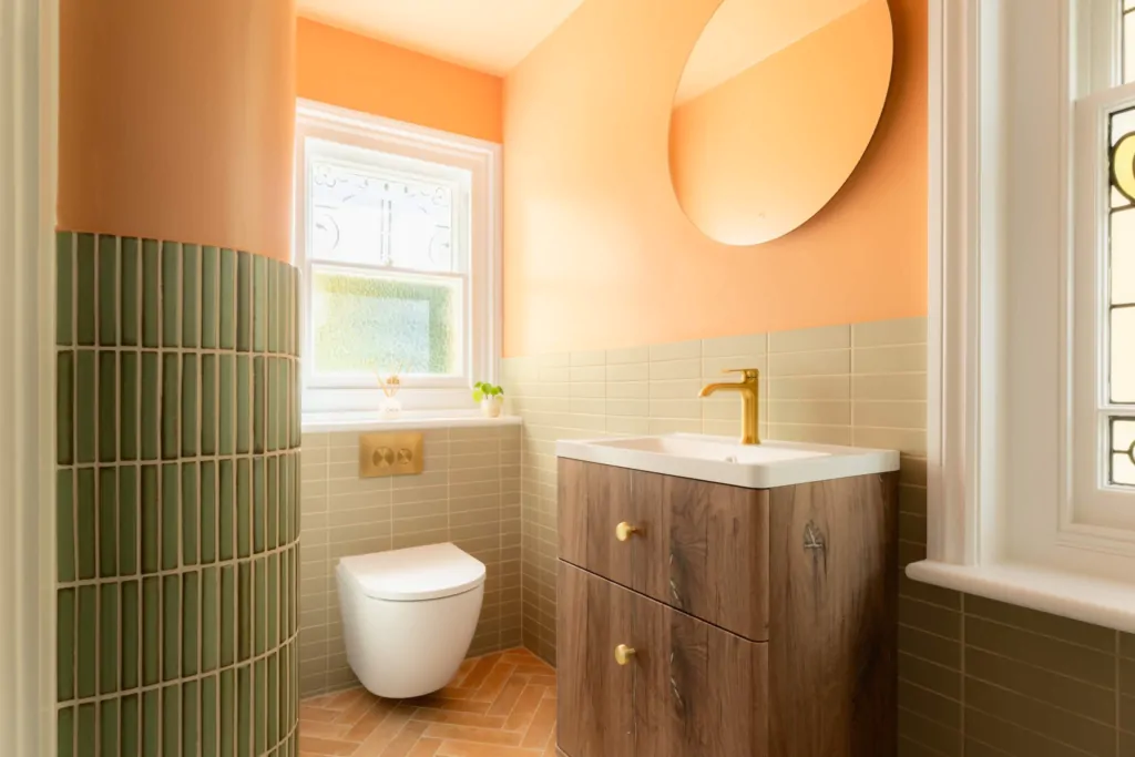 Wide view of the Clementine bathroom showing the curved green tiled partition, wall-hung toilet, wood vanity and terracotta herringbone floor.