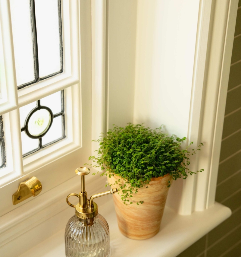 Original stained-glass bathroom window with a potted plant and brass plant mister, complementing the warm green and clementine colour scheme.