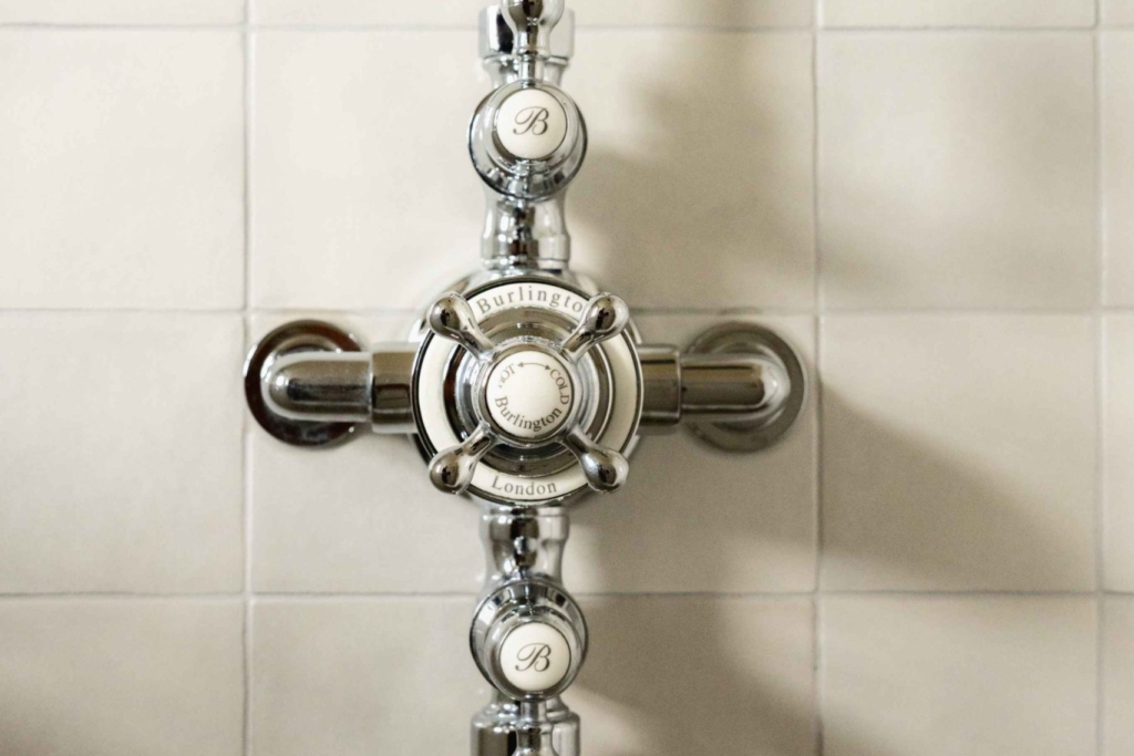 Close-up of traditional Burlington polished chrome shower controls with white ceramic handles against pale marble tiles.