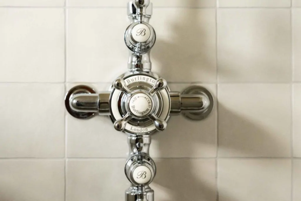 Close-up of traditional Burlington polished chrome shower controls with white ceramic handles against pale marble tiles.