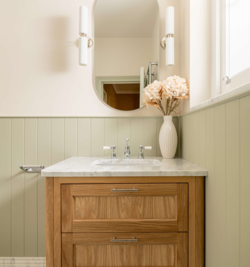 Wall-mounted oak vanity beneath an oval mirror with sage panelling and black-and-white marble basketweave flooring.