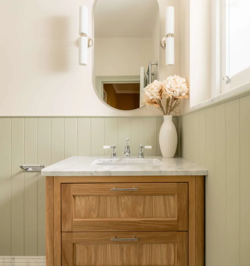 Wall-mounted oak vanity beneath an oval mirror with sage panelling and black-and-white marble basketweave flooring.